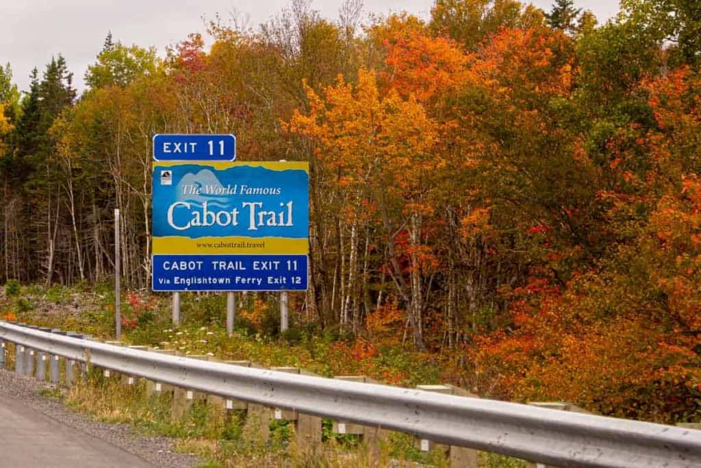 Bright blue and yellow Cabot Trail exit sign on a highway with colorful autumn foliage in the background.