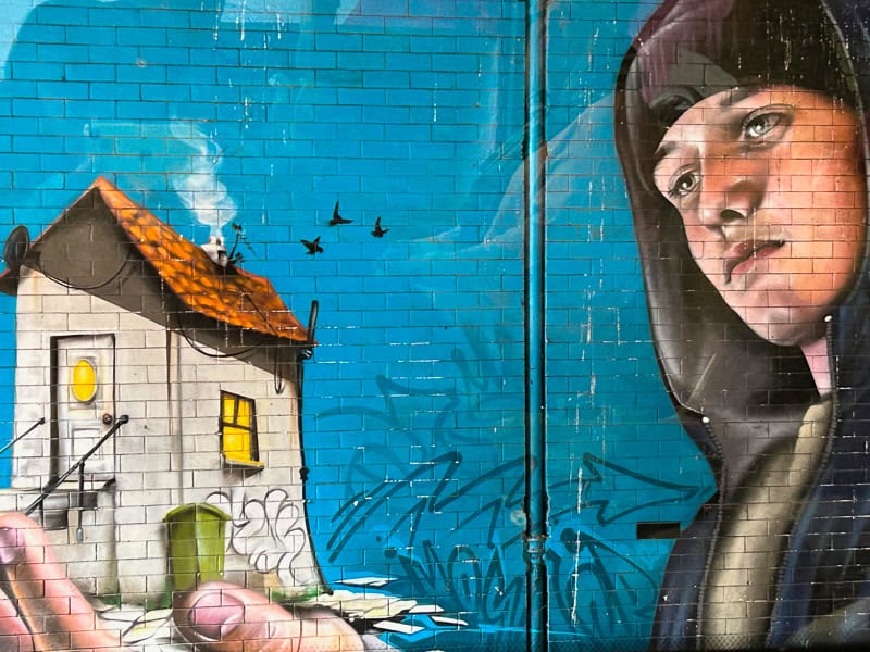 Image on the Glasgow Mural Trail of a young woman in a hoodie cradling a small house in her hand, birds lifting from a chimney, warmth in the windows. 
